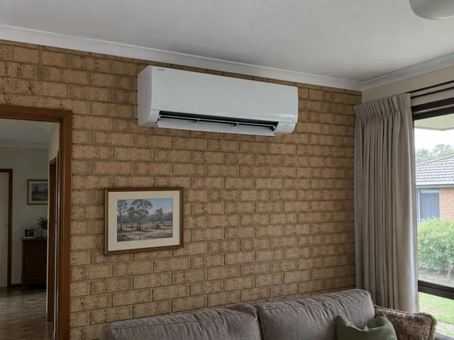 Wall-mounted split system running above a brick feature wall in a living room with curtains in Andrews Farm