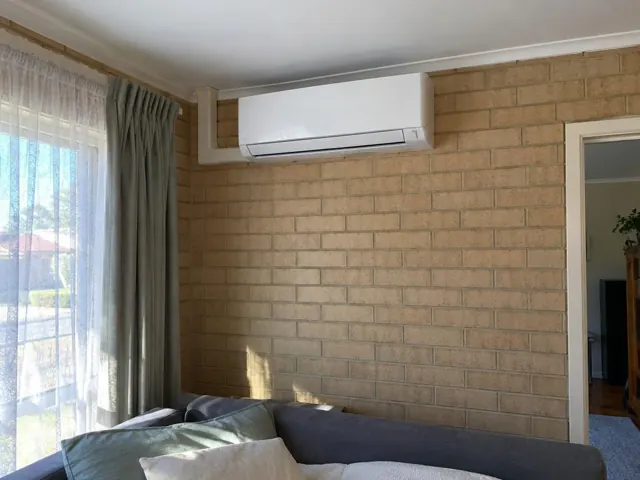 Wall-mounted split system indoor unit on a brick wall above a couch and curtains in a living room in Davoren Park